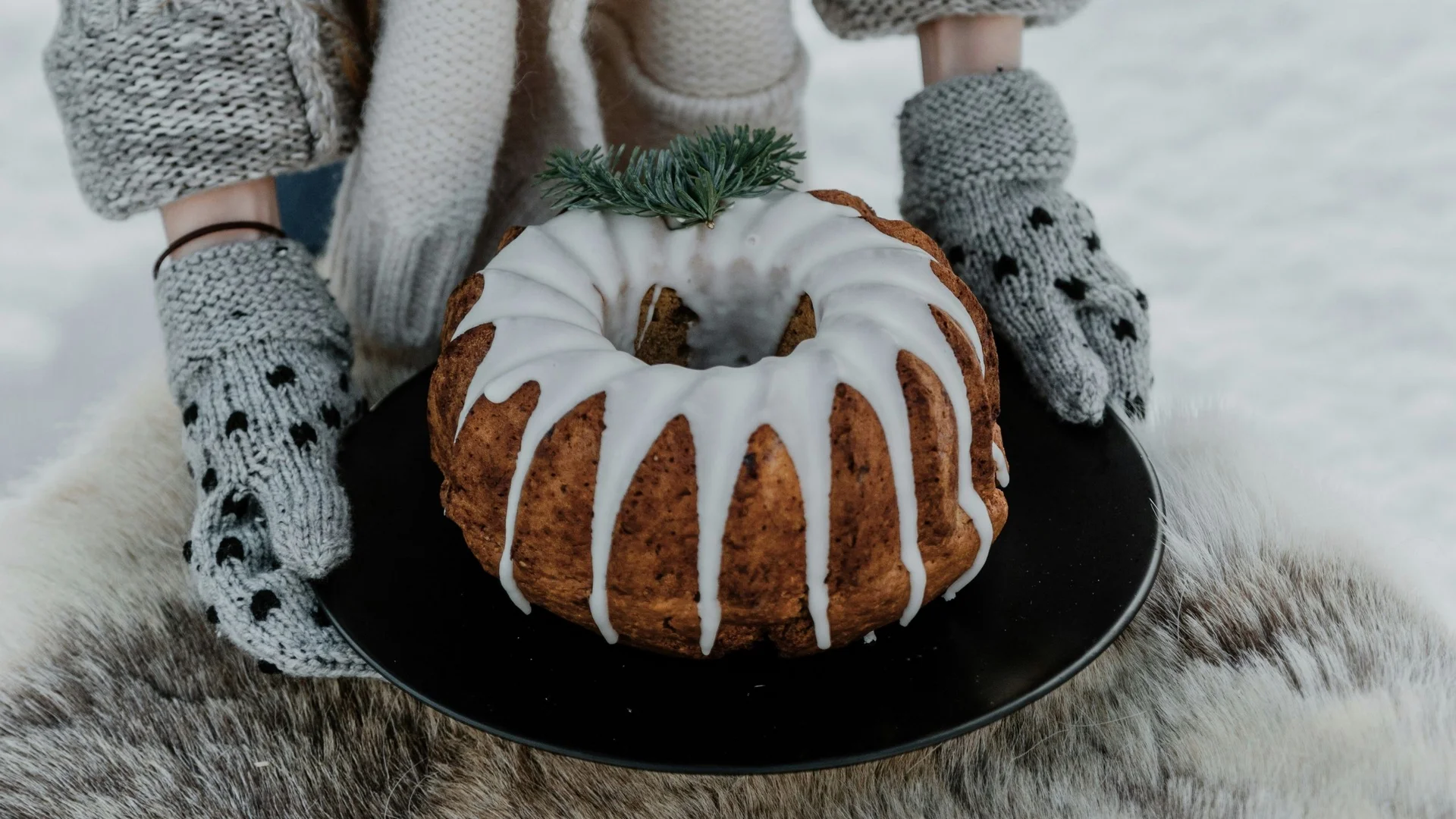 Eierlikör-Gugelhupf Person hält Kuchen mit Zuckerguss auf schwarzem Teller, trägt graue Strickhandschuhe