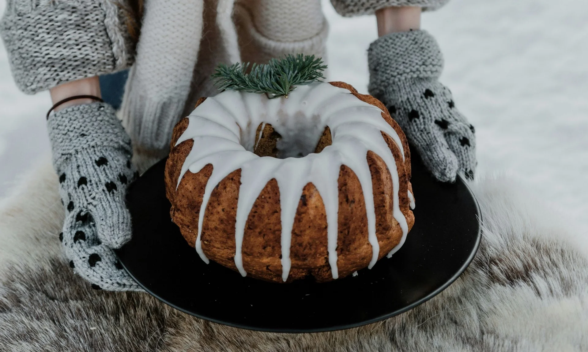 Geburtstagspost Person hält Kuchen mit Zuckerguss auf schwarzem Teller, trägt graue Strickhandschuhe
