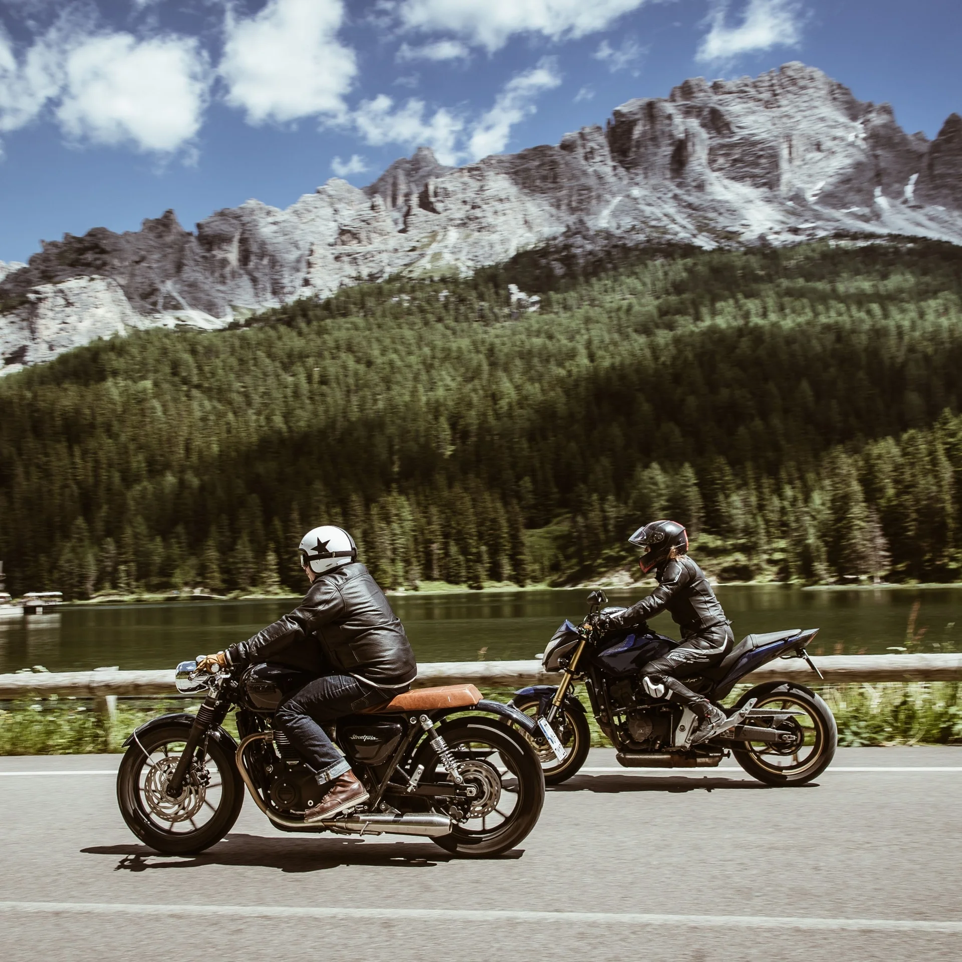 Zwei Motorradfahrer fahren vor Berglandschaft und Wald auf Straße entlang See