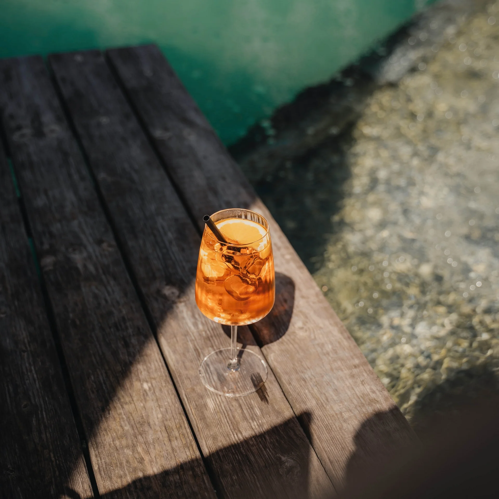 A glass of Aperol Spritz with ice and orange slice on wooden deck by the water