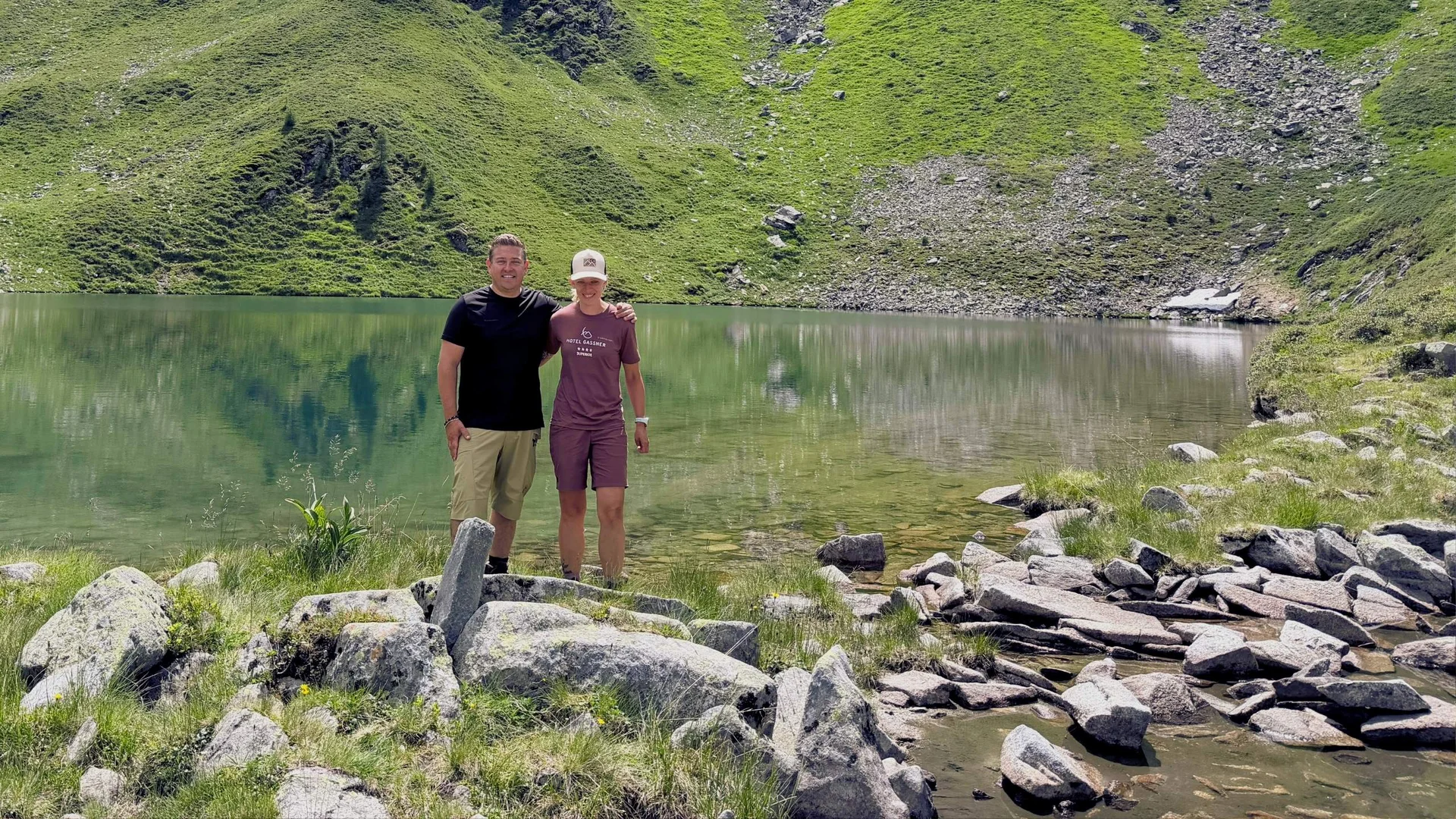 Hiking Service Two people standing by a mountain lake with green hills in the background