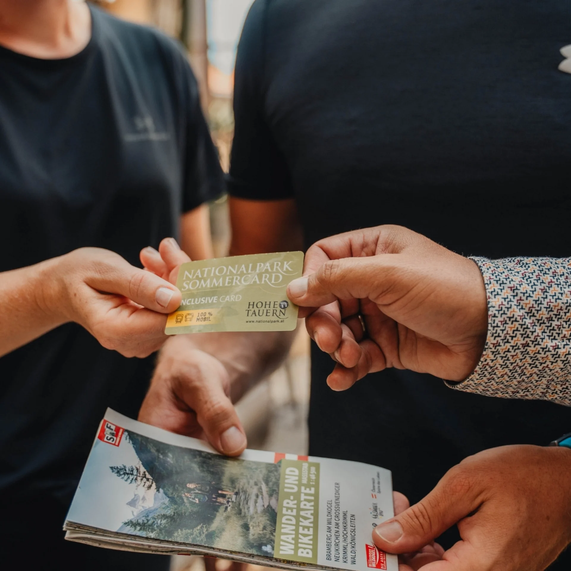 Person übergibt Nationalpark Sommercard an anderen, hält Wander- und Bikerkarte