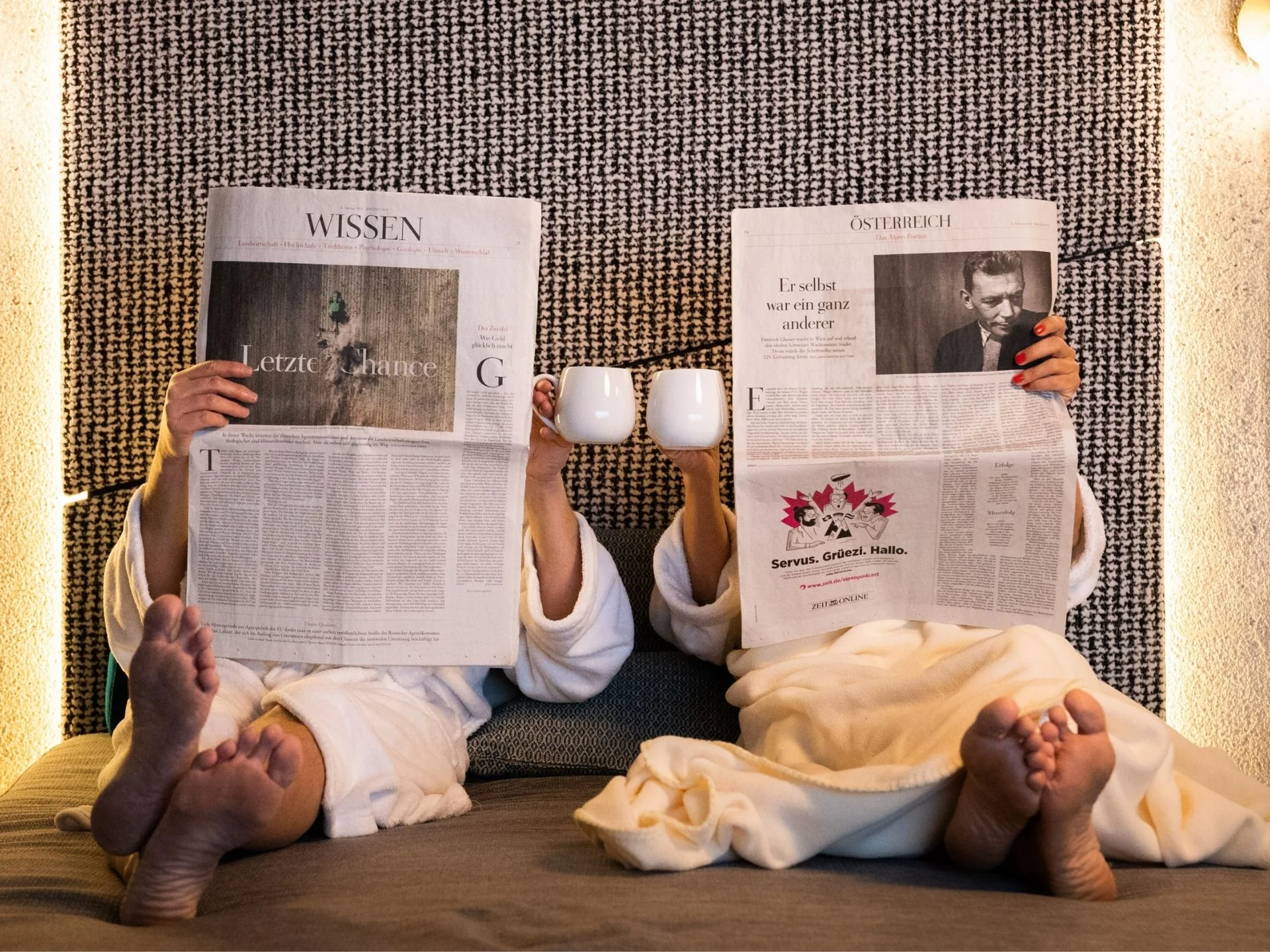 Zwei Personen in Bademänteln lesen Zeitung und stoßen mit Tassen an