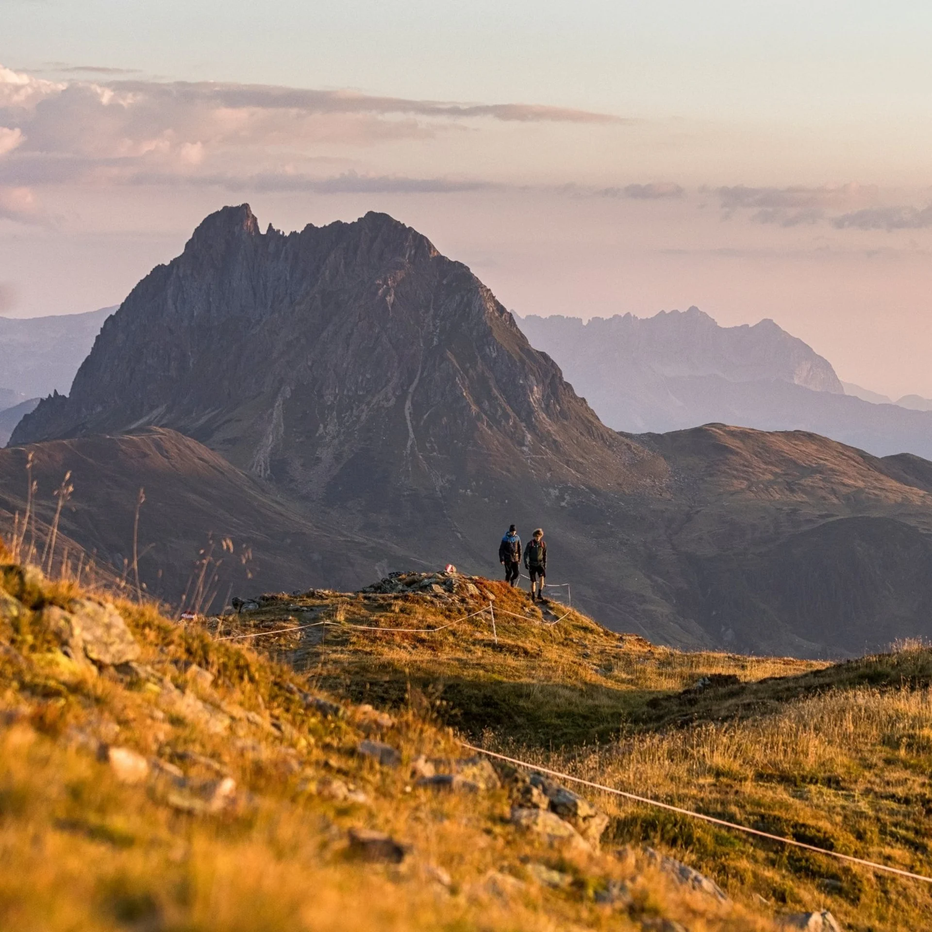 Hikers on a trail overlooking a prominent mountain at sunset