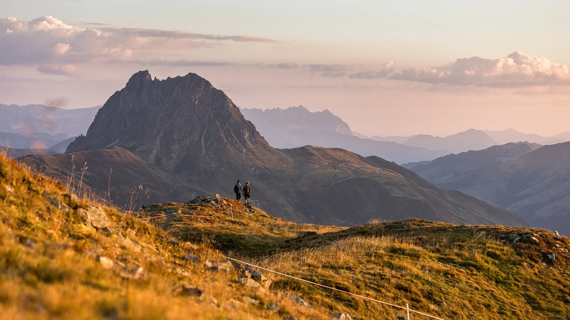 Hikers on a trail overlooking a prominent mountain at sunset