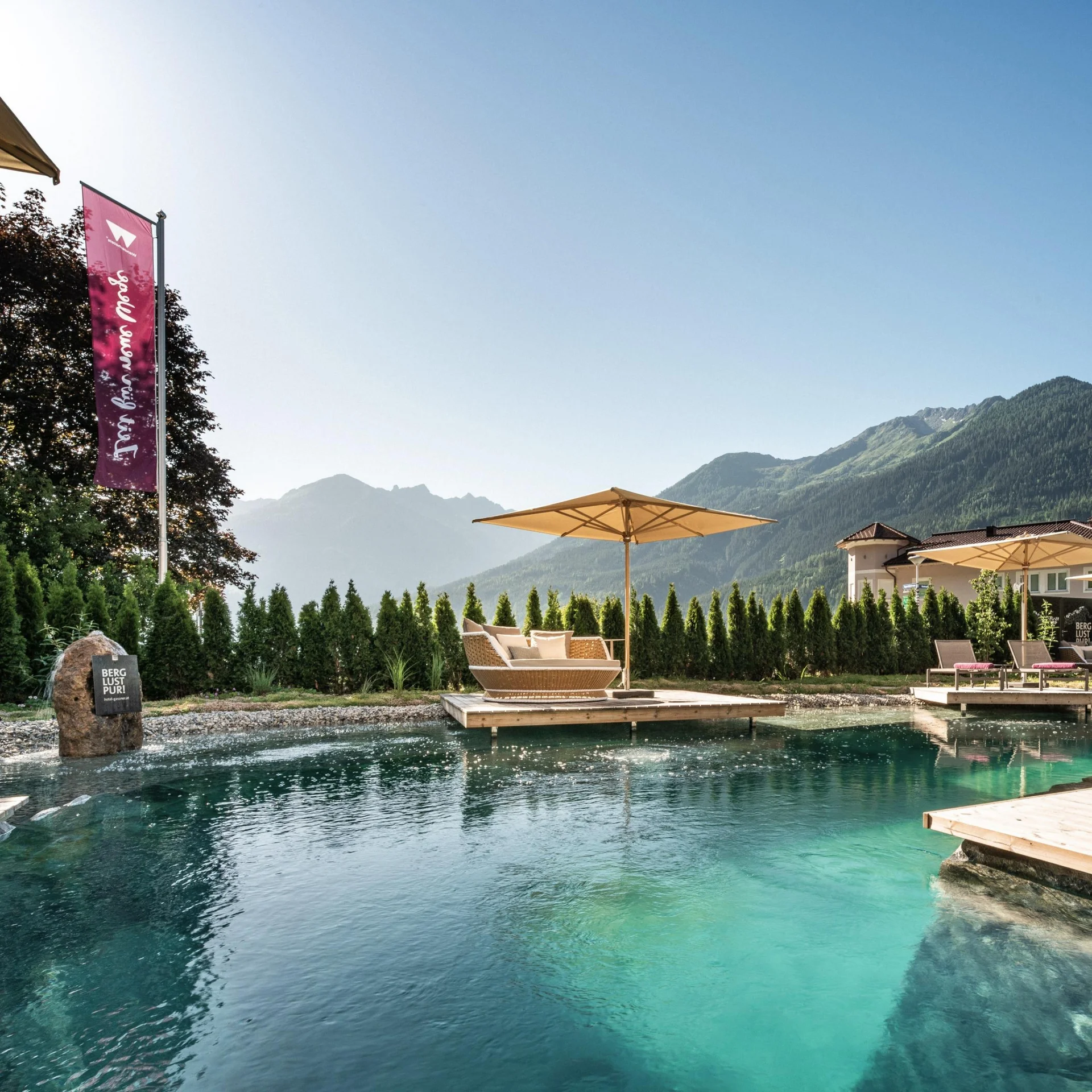 Feel.Good Natürlicher Pool mit Sonnenliegen und Bergpanorama unter klarem Himmel