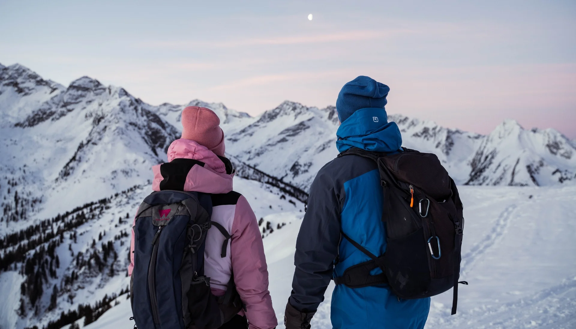 Two hikers in winter gear looking at snow-covered mountains at dusk