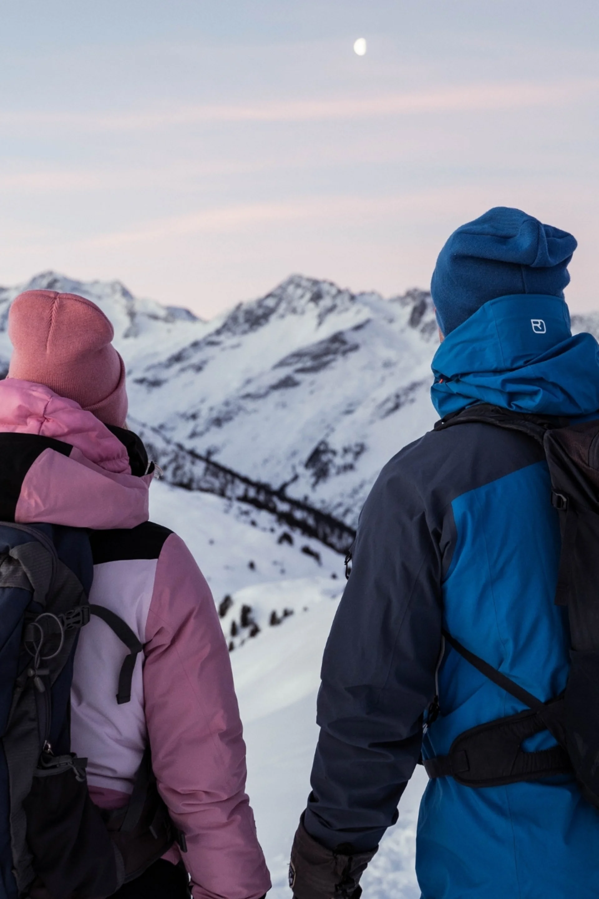 Zwei Wanderer in Winterkleidung blicken auf verschneite Berge bei Dämmerung