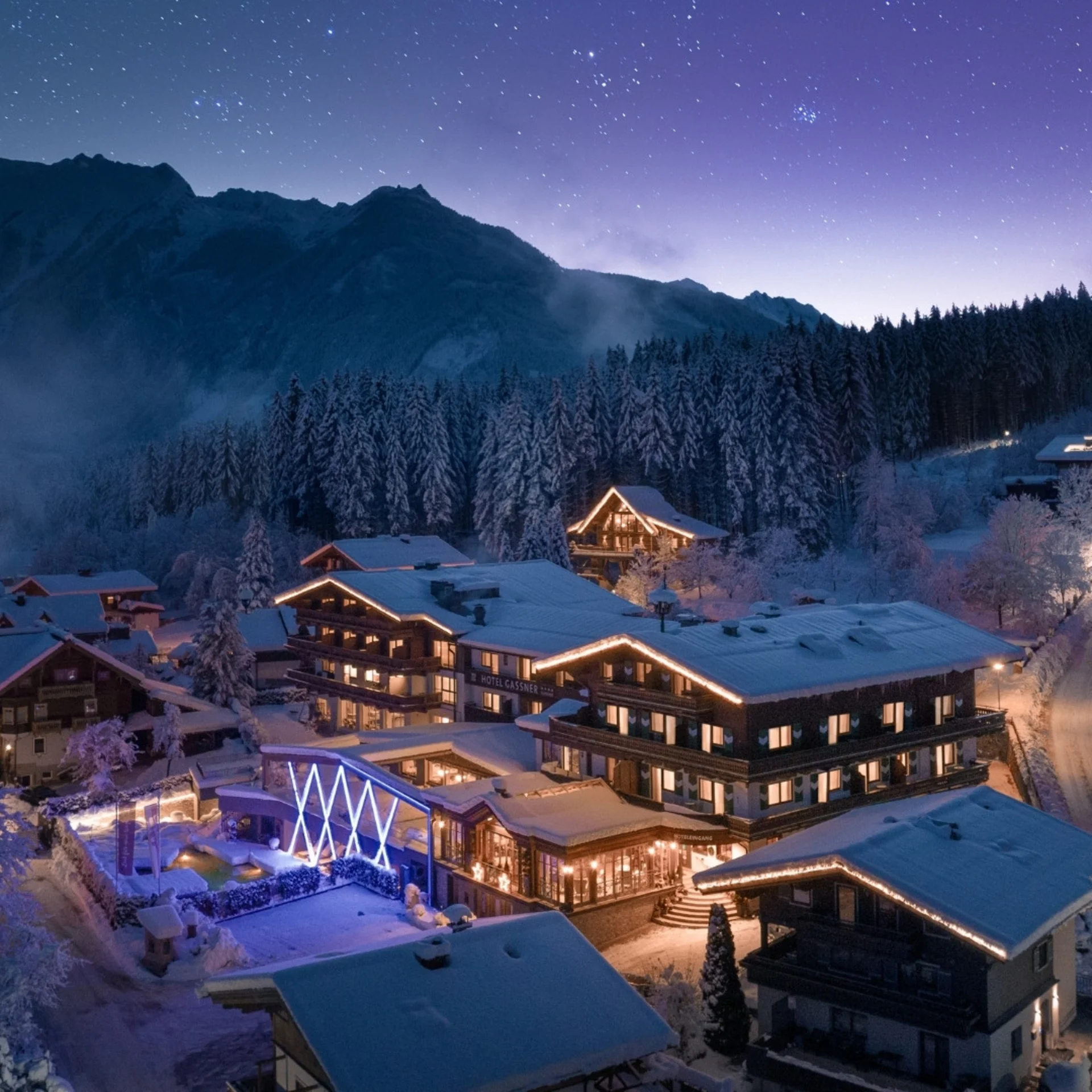 Christ.Kindl Verschneites Bergdorf bei Nacht mit beleuchtetem Hotel, dunklen Bergen und sternenklarem Himmel