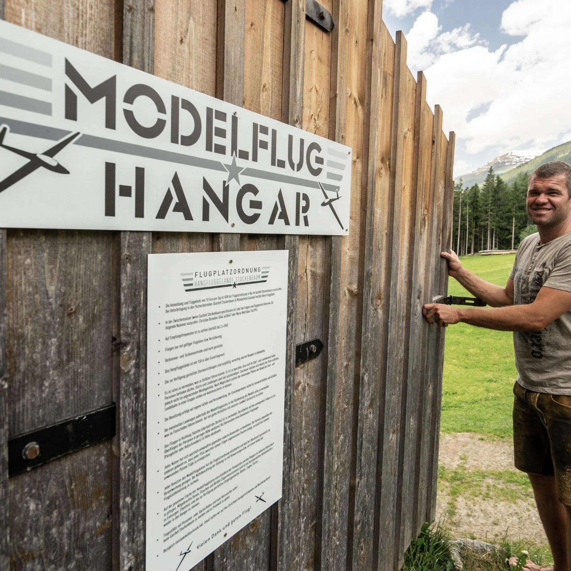 Man opening wooden gate to model airplane hangar in green mountain landscape