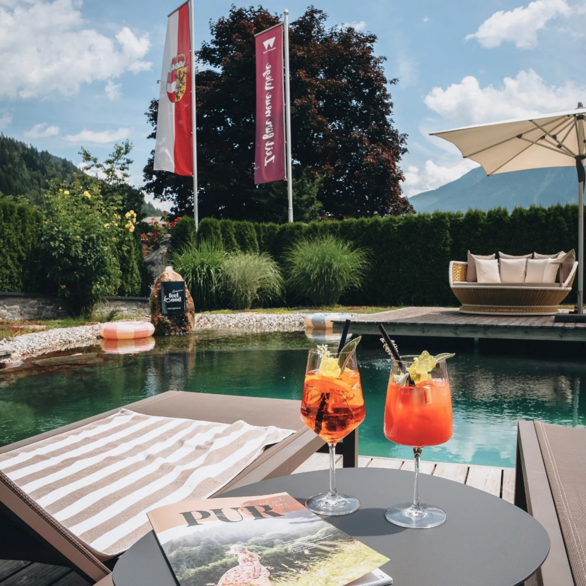 Zwei Cocktails auf Tisch neben Liegestuhl am Pool mit Sonnenschirm und Garten