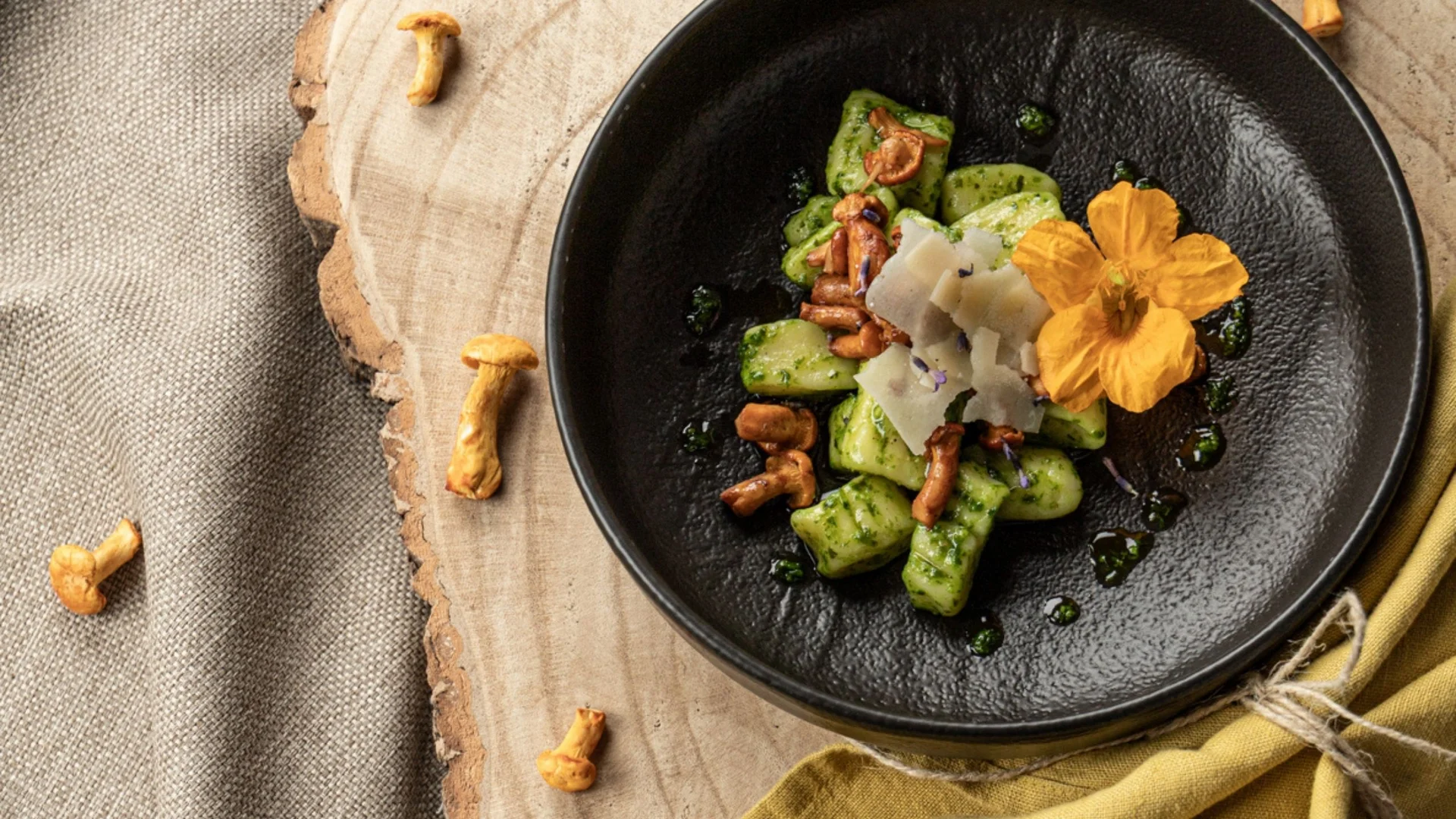 Gregors Sommerrezept: Gnocchi mit Basilikumpesto und Eierschwammerln Schwarzer Teller mit grünen Gnocchi, Pilzen, Käse und essbarer Blume auf Holzbrett
