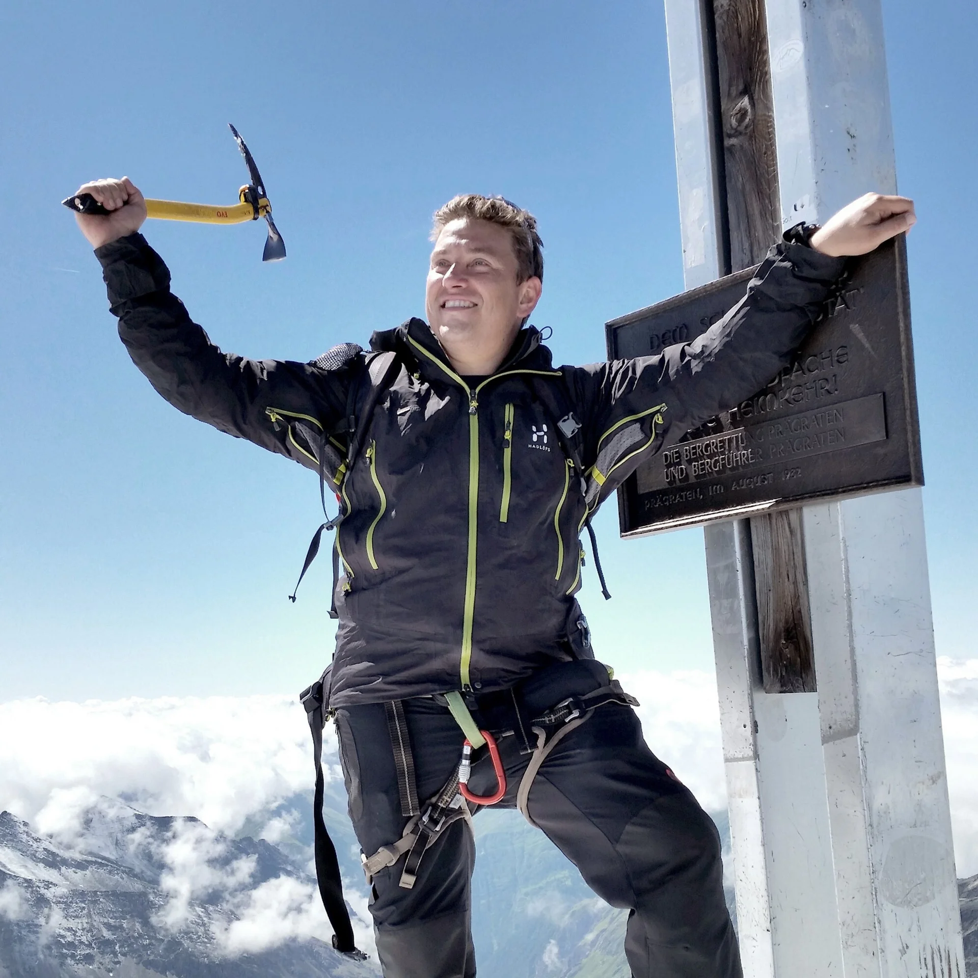 Glücklicher Bergsteiger mit Eispickel am Gipfelkreuz auf bergigem Hintergrund