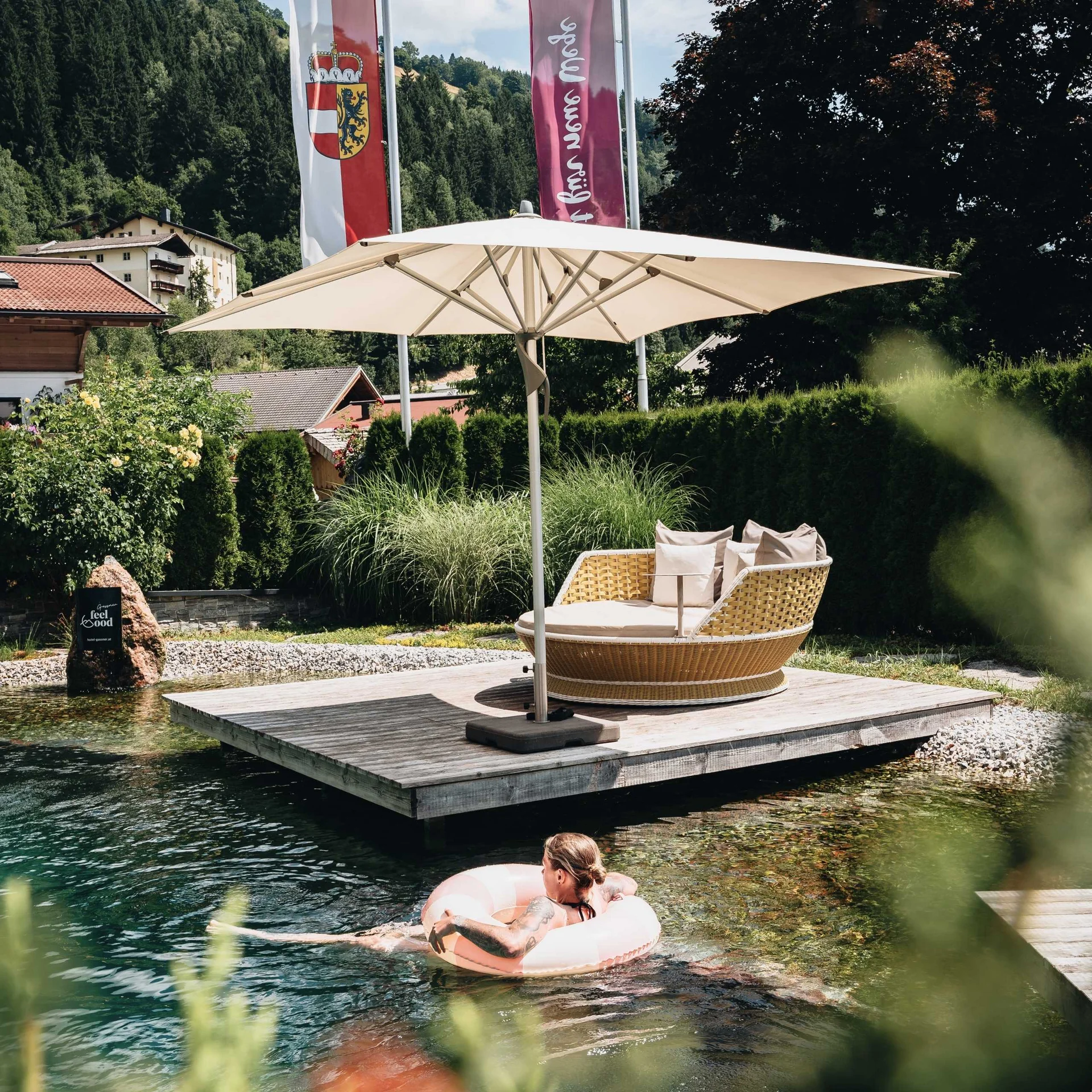 Person schwimmt mit Schwimmring in klarem Naturteich neben Liegefläche und Sonnenschirm