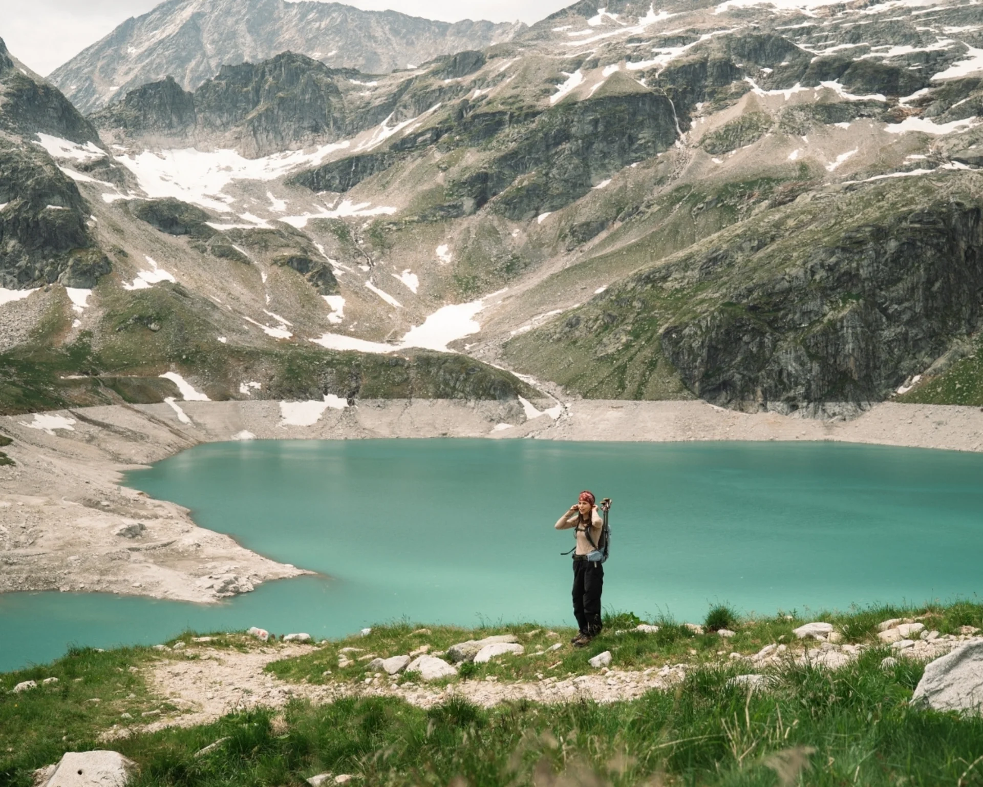 Wanderer steht am Ufer eines türkisfarbenen Bergsees mit schneebedeckten Gipfeln