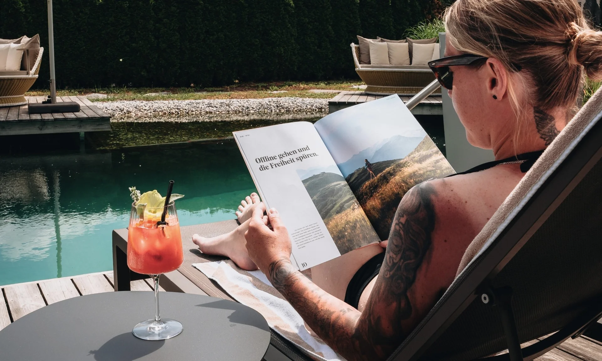 Person liest Buch und entspannt am Wasser mit Cocktail auf Tisch
