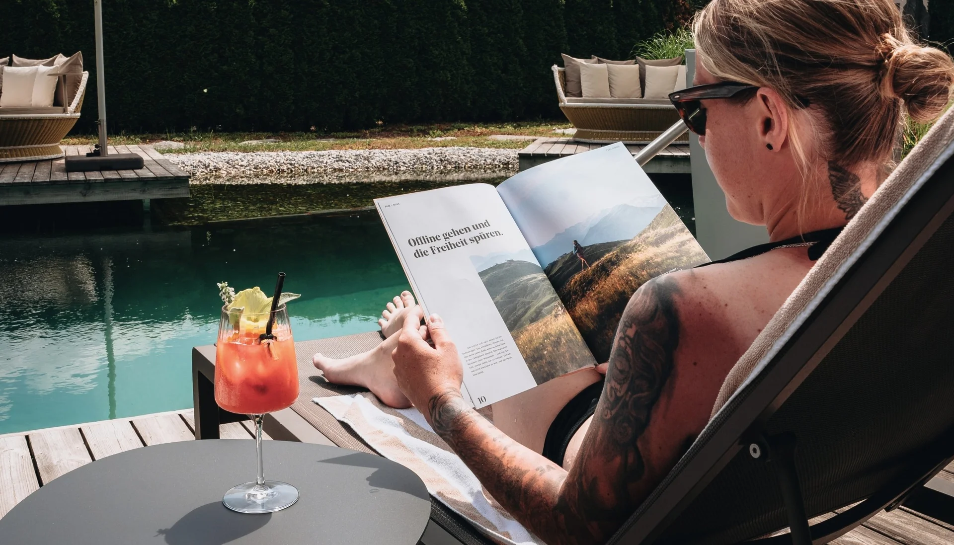 Person liest Buch und entspannt am Wasser mit Cocktail auf Tisch