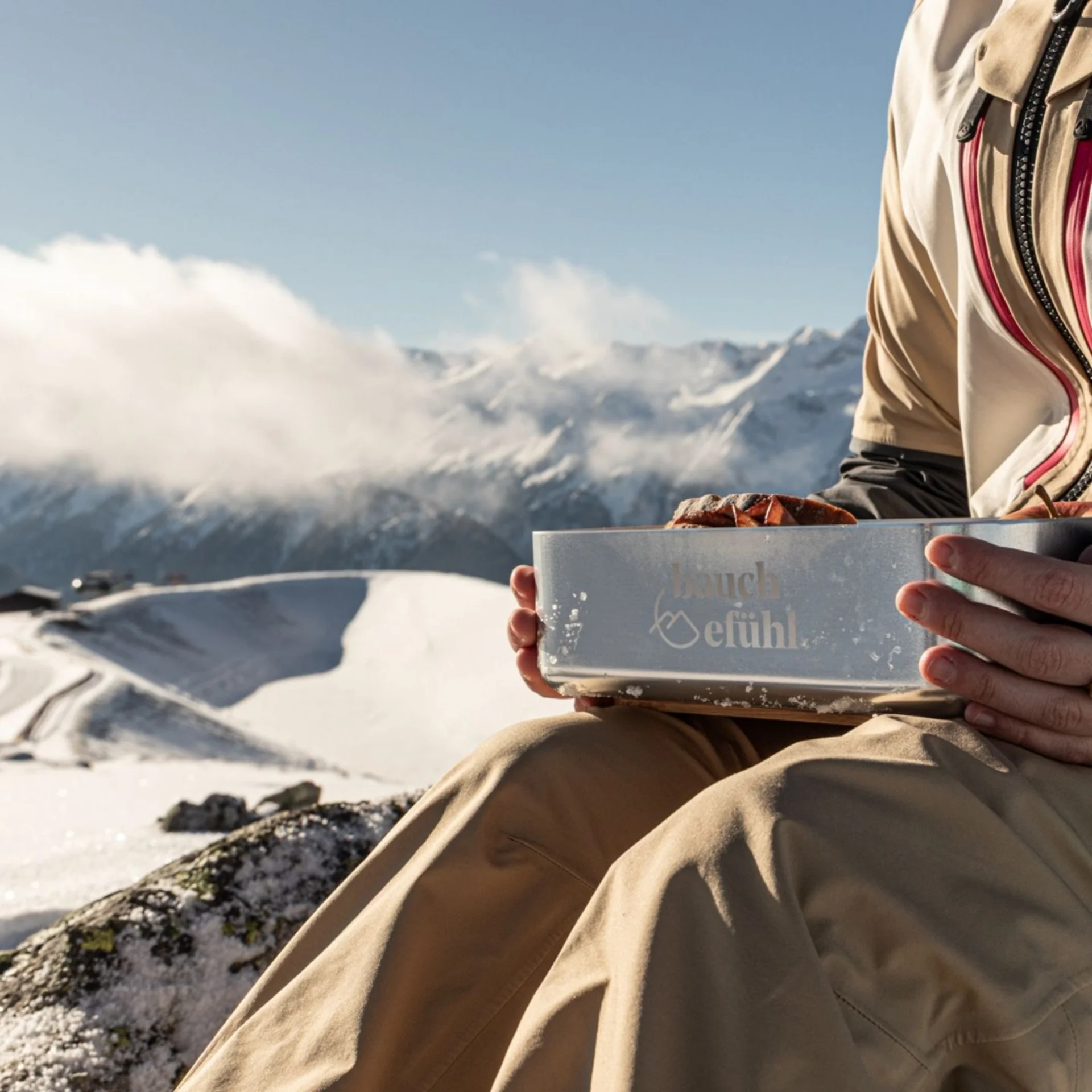 Person sitzt auf verschneitem Berg mit Lunchbox in der Hand