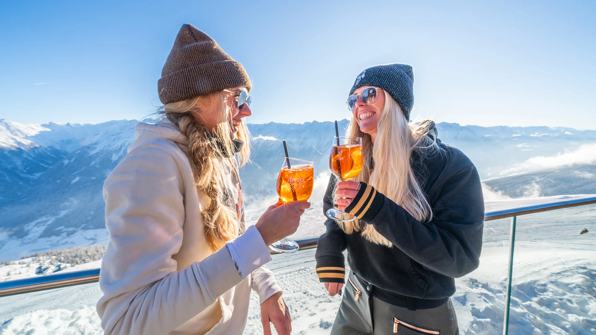 Winterkinder unter sich Zwei Frauen genießen Aperol Spritz auf einer sonnigen Bergterrasse im Schnee