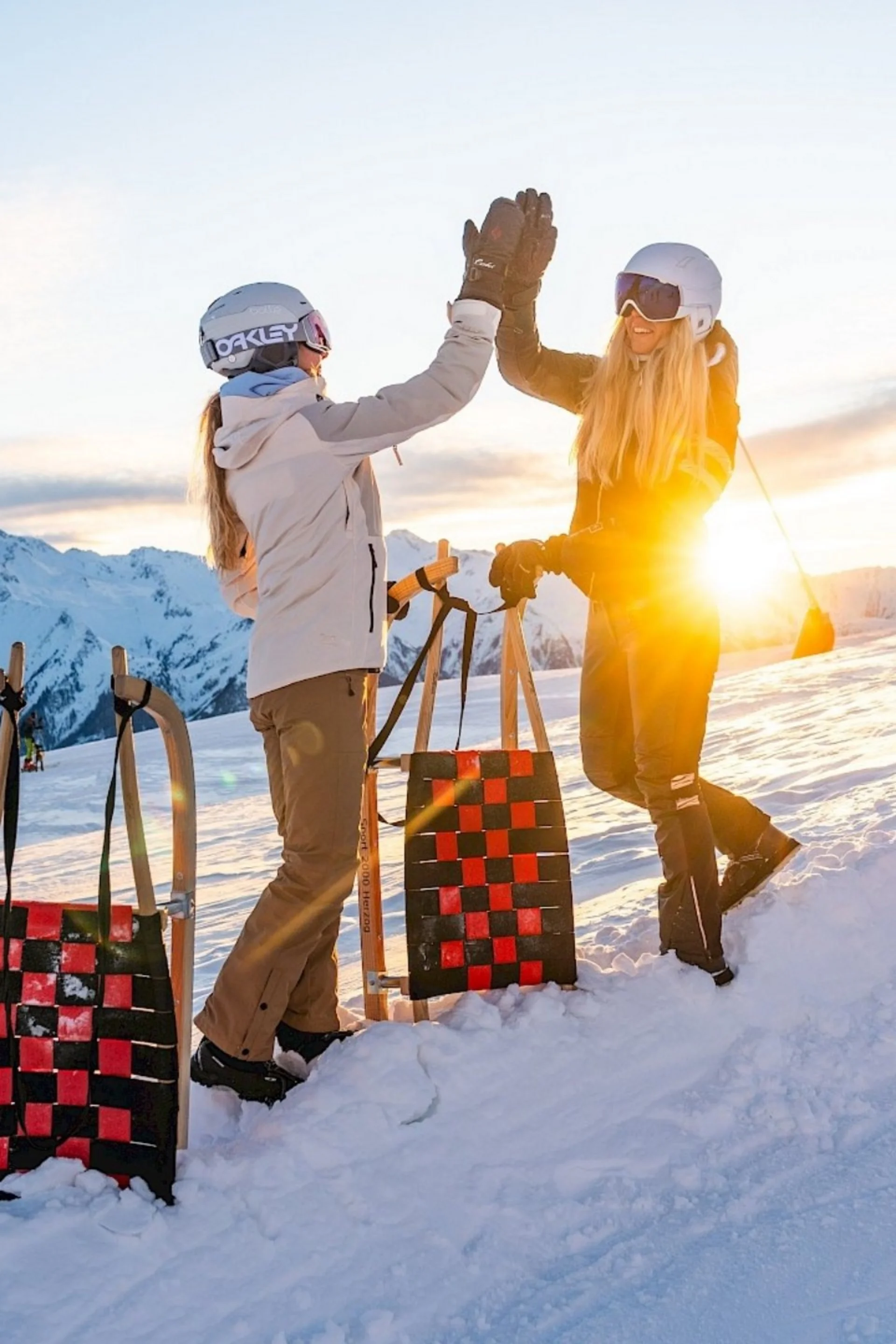 Zwei Personen auf verschneitem Berg geben sich High Five mit Schlitten im Sonnenuntergang