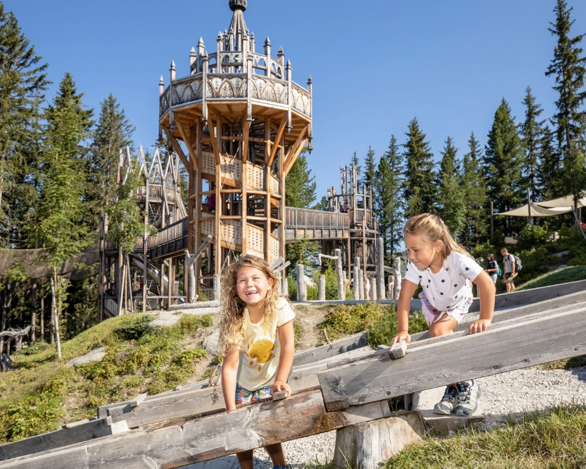 Zwei Mädchen spielen auf einem Holzspielplatz mit Turm im Wald bei Sonnenschein