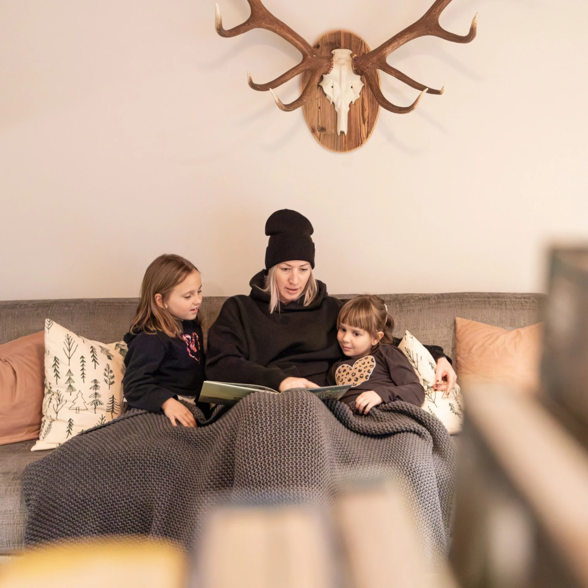 Frau liest mit zwei Kindern auf dem Sofa unter einer Decke vor, Hirschgeweih an der Wand