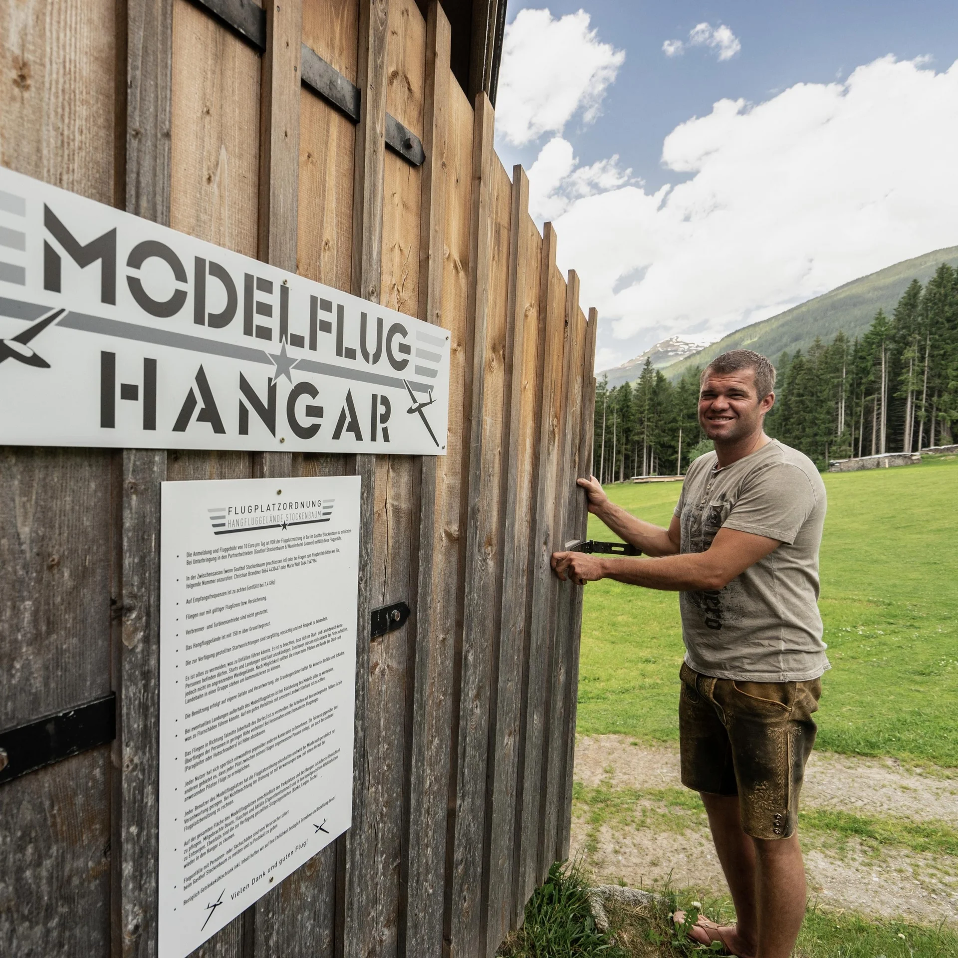 Lächelnder Mann öffnet Holztür zu Modellflughangar in Berglandschaft