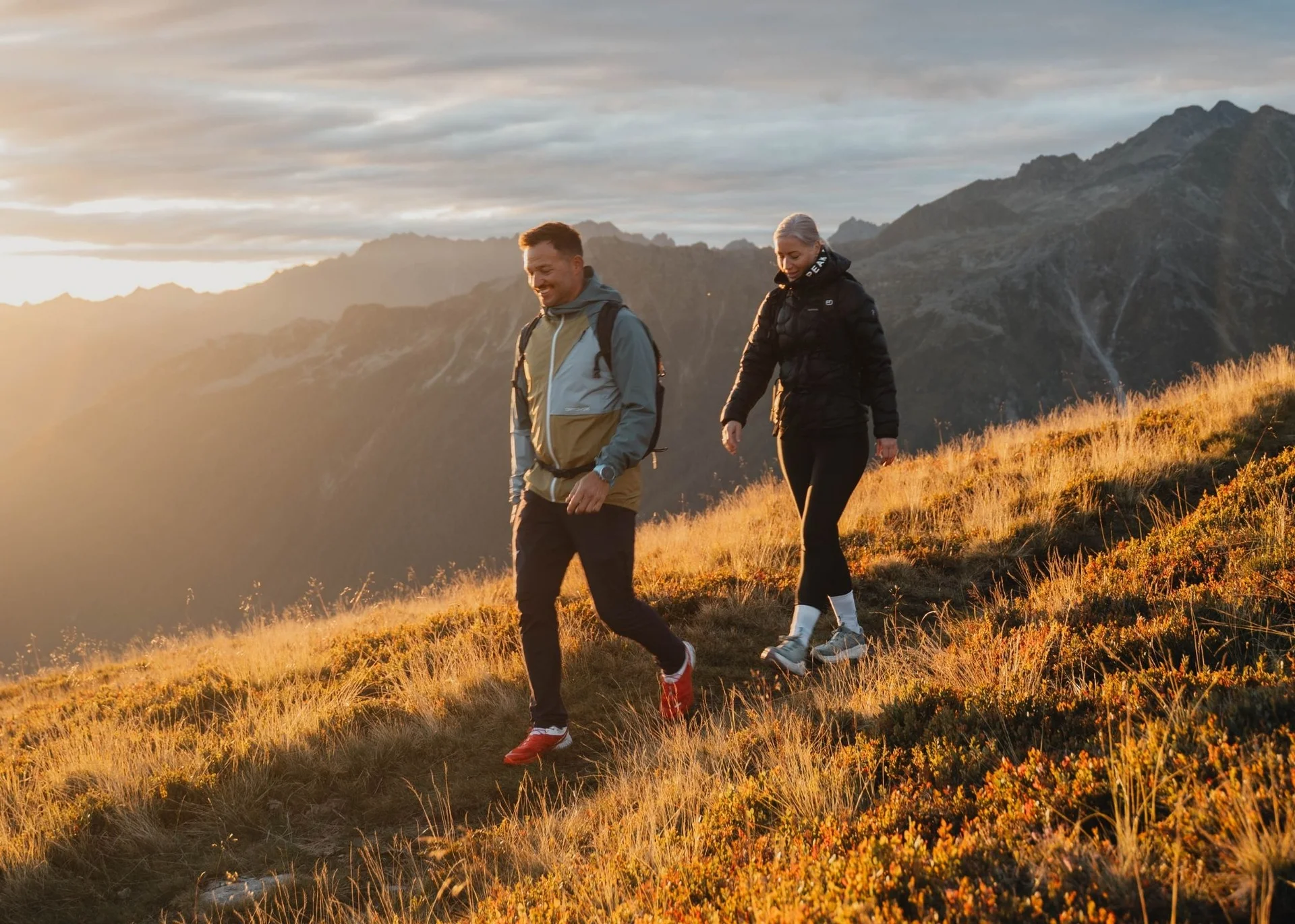 Home Paar wandert bei Sonnenuntergang auf einem grasbewachsenen Bergpfad mit Bergen im Hintergrund