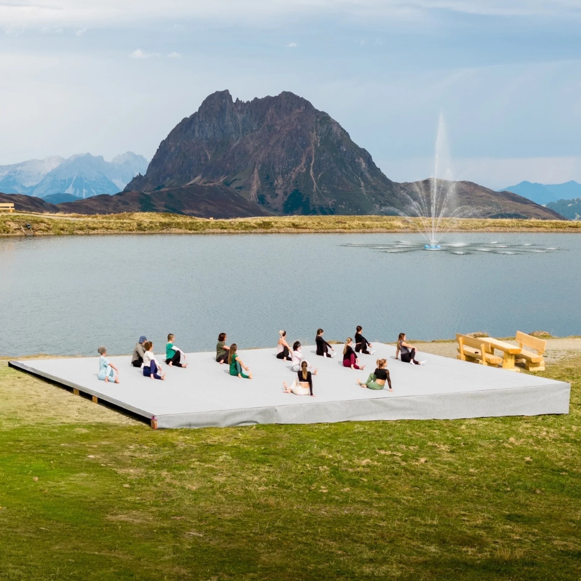 Menschen praktizieren Yoga auf einer Plattform vor einem Bergsee mit Springbrunnen