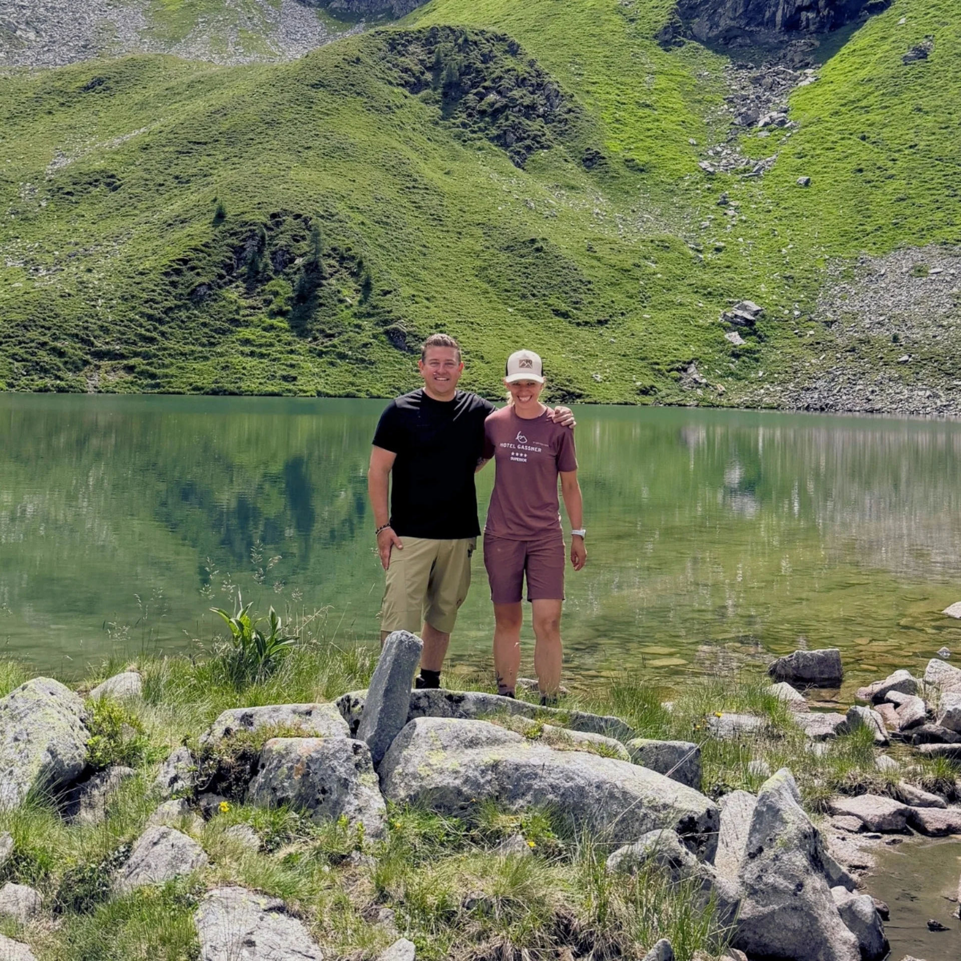 Zwei Personen stehen am Ufer eines Bergsees mit grünen Hügeln im Hintergrund